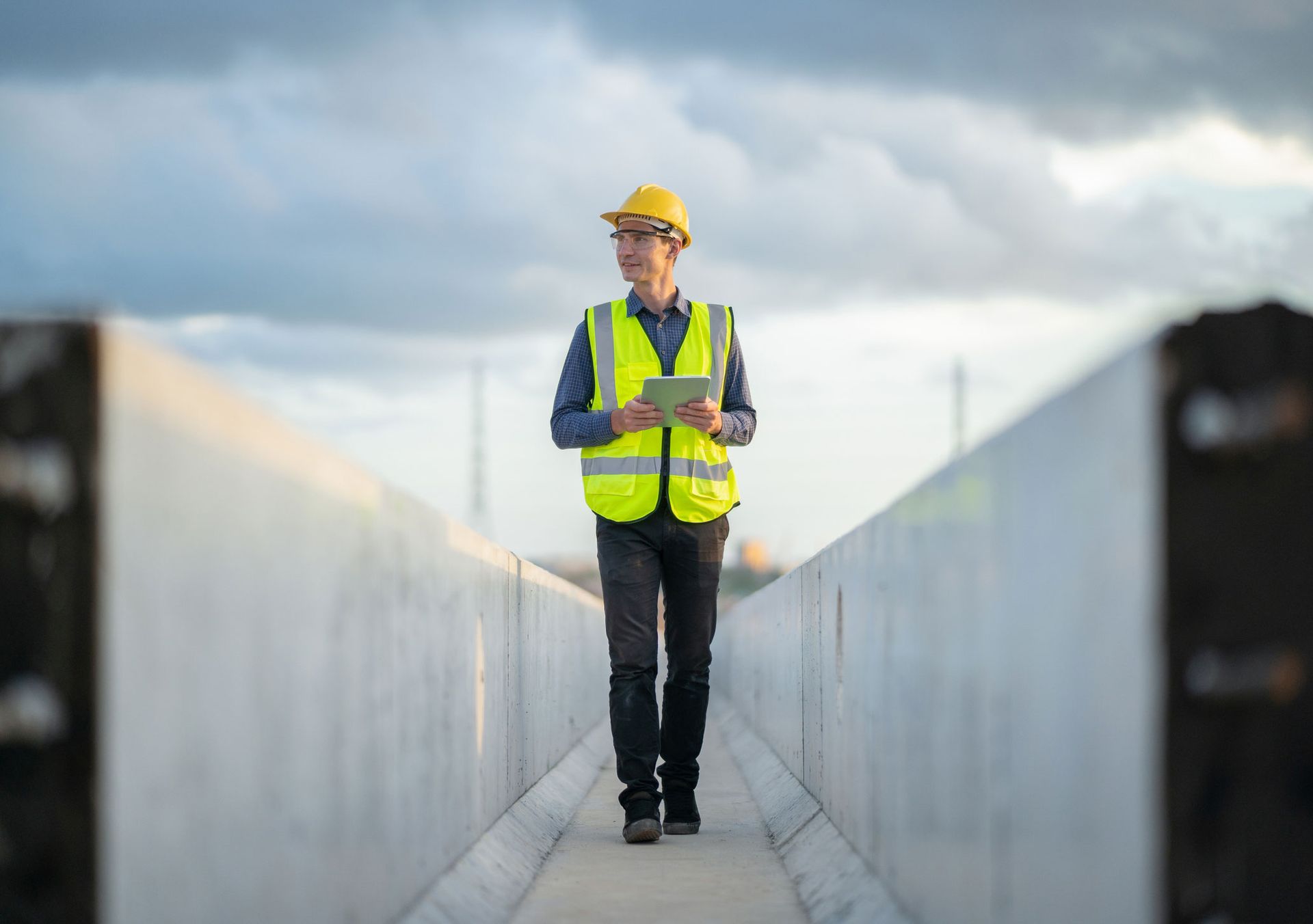 Construction worker in a yellow vest and helmet holding a tablet on a site.