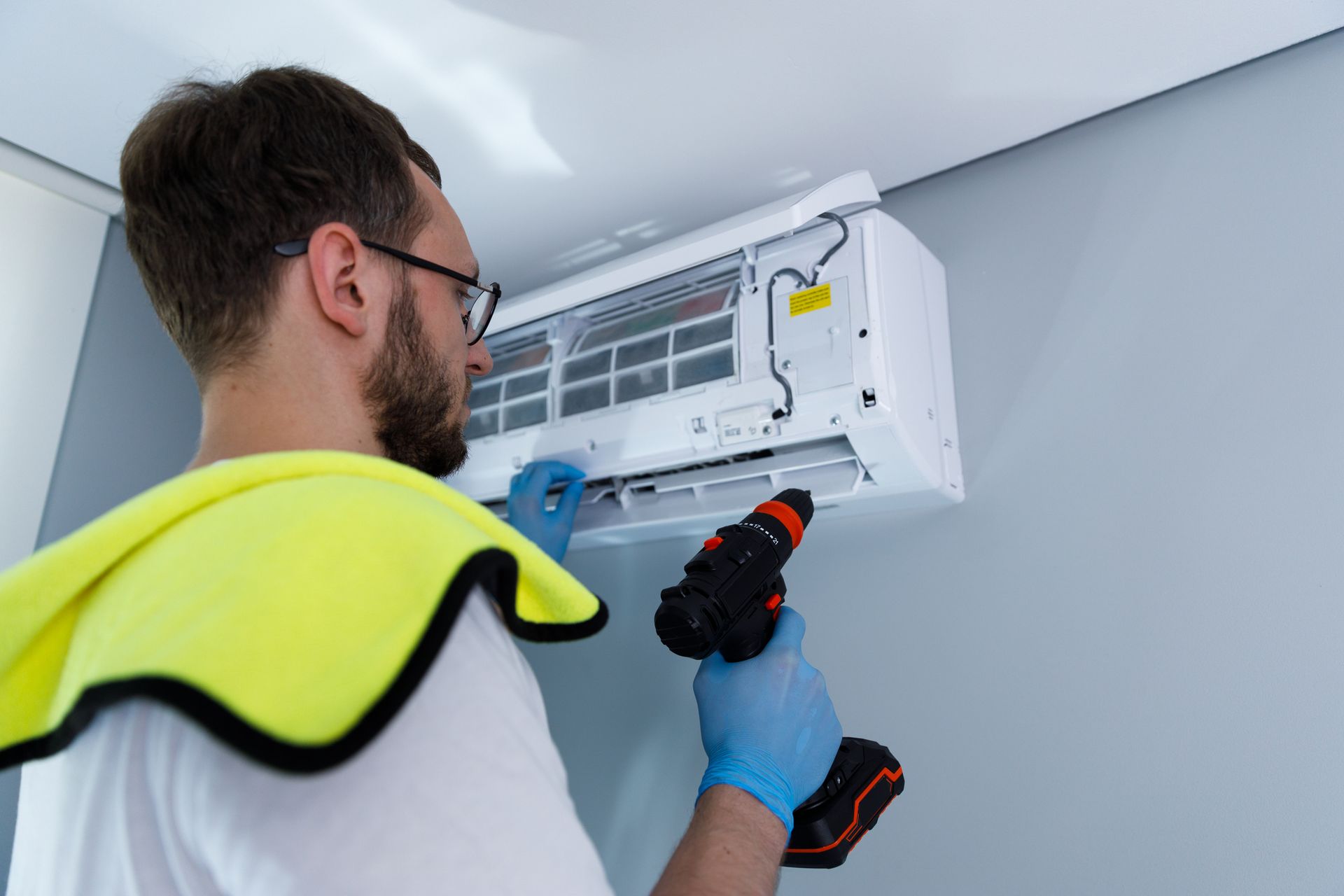Person in blue gloves using a drill to repair a white air conditioner on a gray wall.