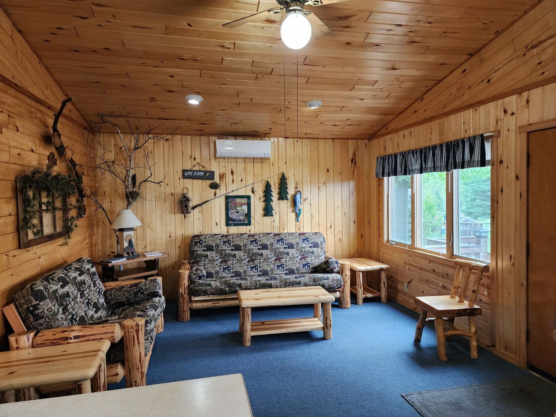 Wood-paneled rustic living room with blue carpet, floral sofas, and a windowed wall.