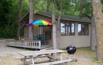 Cabin with porch, rainbow umbrella, picnic table, and charcoal grill in a wooded campsite