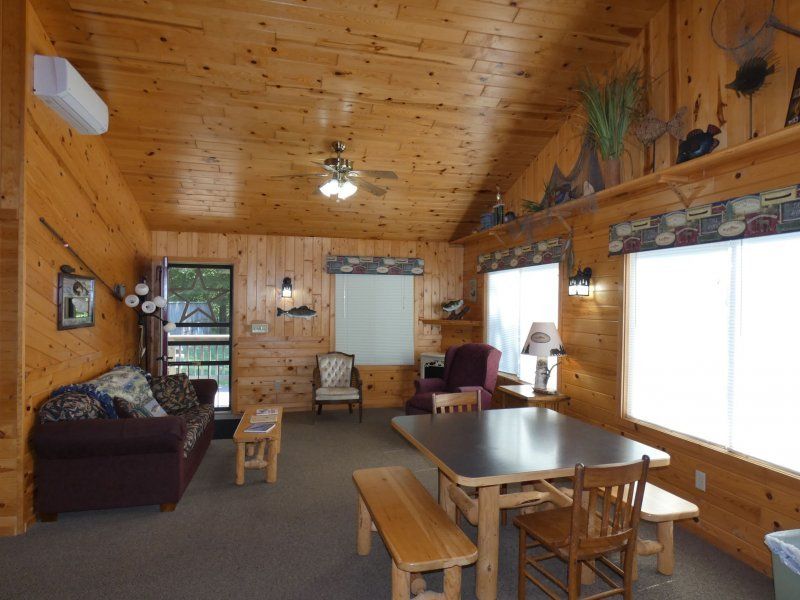 Cozy wood-paneled cabin living room with sofa, dining table, chairs, and large windows