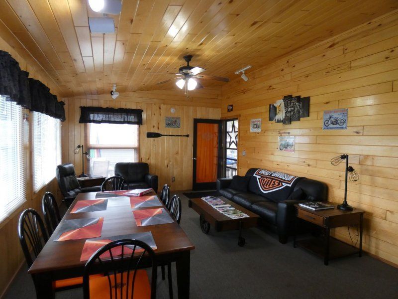 Cozy wood-paneled living room with a long dining table, sofa, coffee table, and windows with curtains