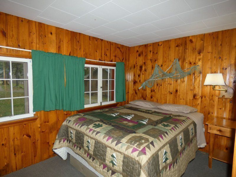 Wood-paneled bedroom with a bed, green curtains, windows, and a lamp by the wall