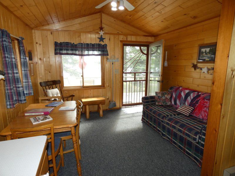 Wood-paneled cabin living room with plaid sofa, small table, window, and open door to a porch