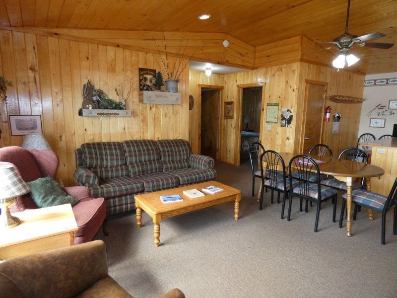 Cozy wood-paneled living room with plaid sofa, armchairs, coffee table, and dining nook.