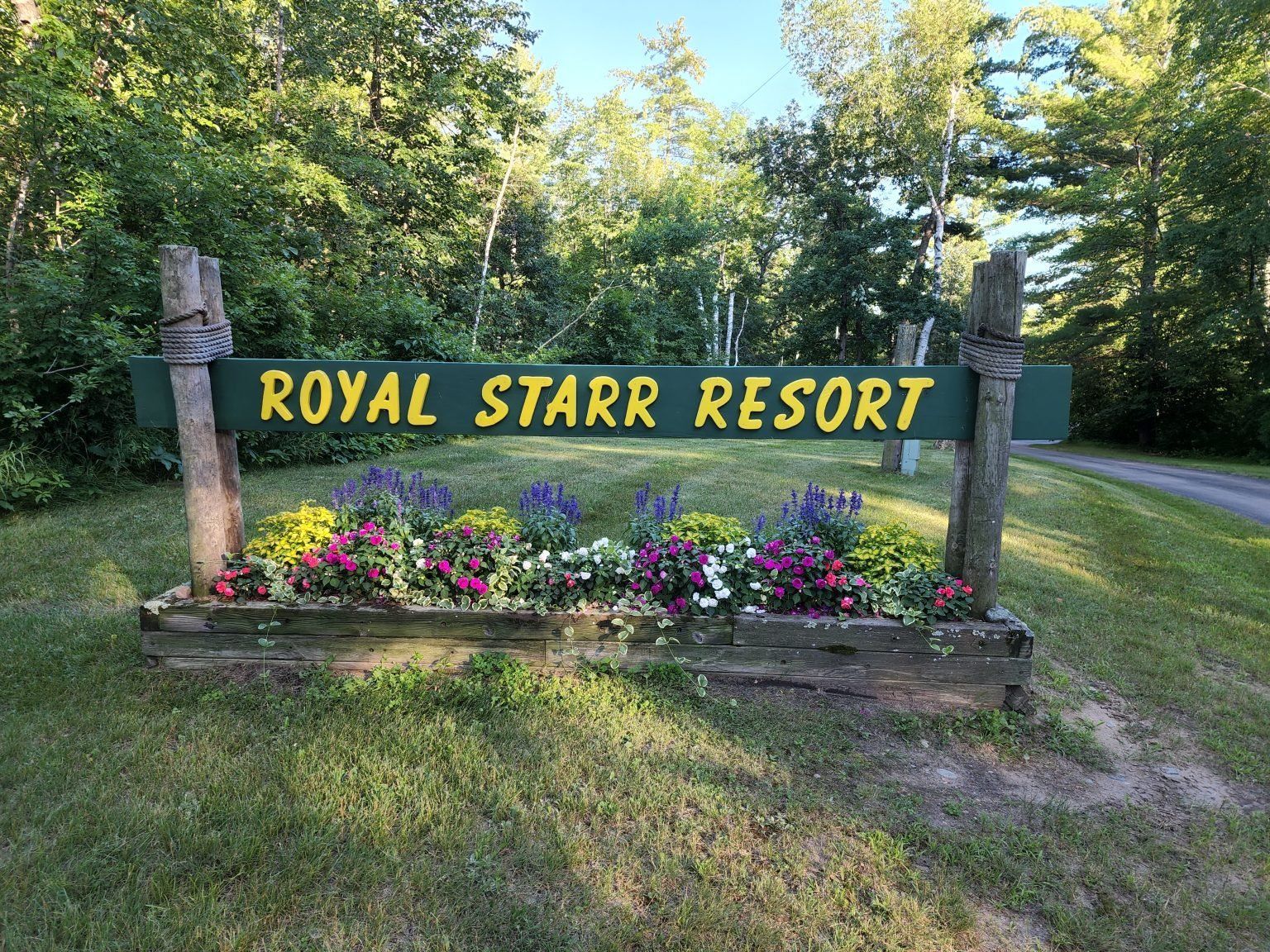 Royal Starr Resort wooden sign with flowers in a sunny wooded roadside setting