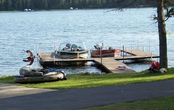 Dock with boats on a lake beside a grassy shore and paved road, with a person near the left boat