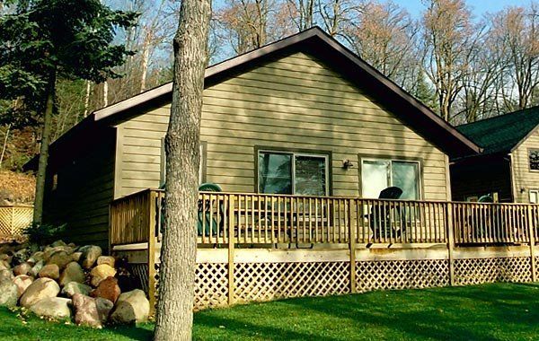 Small beige cabin with a wooden deck, surrounded by trees and rocks in a wooded yard.