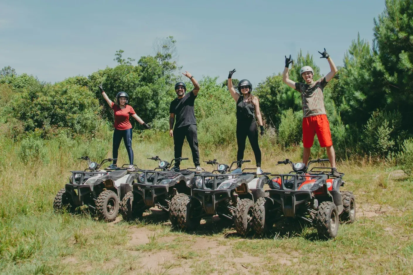 Four people posing on rugged ATVs in a grassy field, arms raised in celebration, with trees in the background