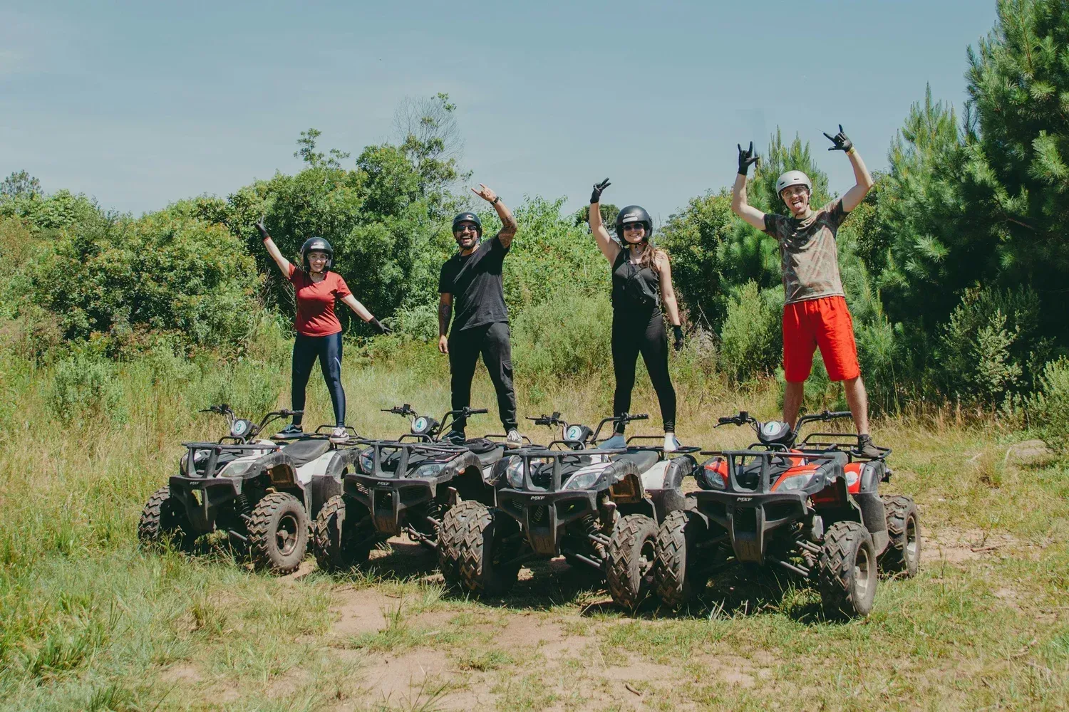 Four people posing on rugged ATVs in a grassy field, arms raised in celebration, with trees in the background