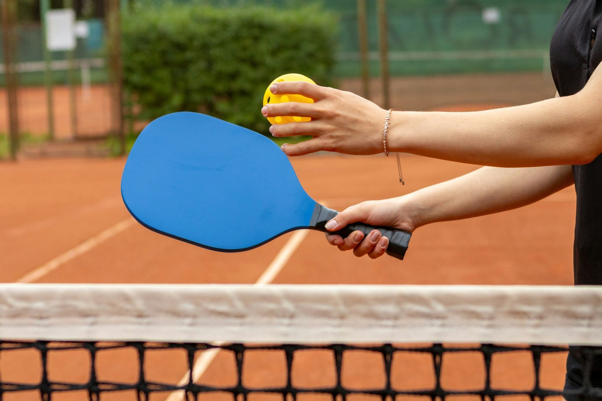 Hands holding blue and black pickleball paddles at a court net with yellow ball in background