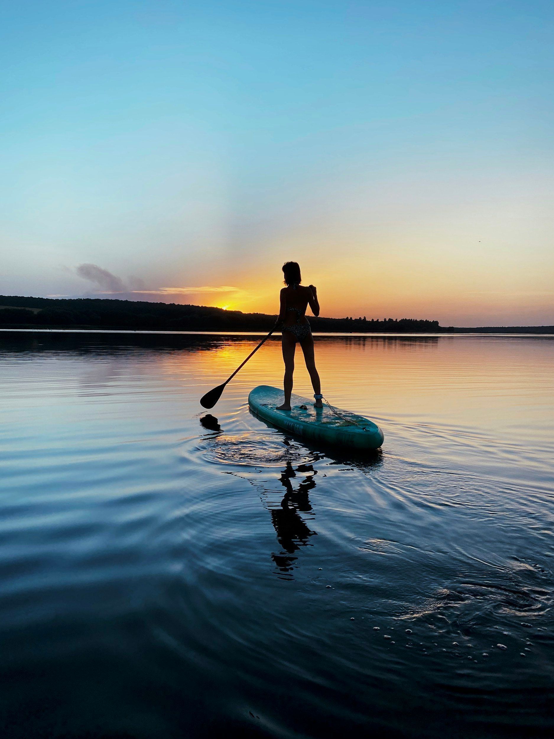 Silhouette standing on a paddleboard at sunset, paddling across calm water with a colorful sky reflection.