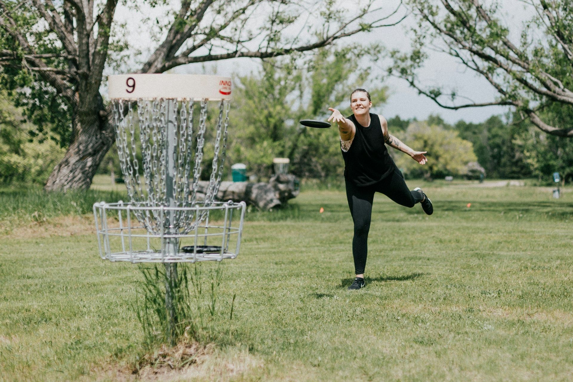 Person mid-throw at an outdoor disc golf basket in a grassy park.