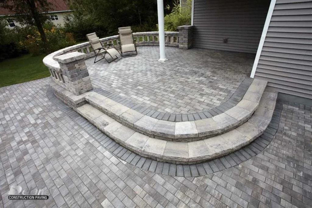 Curved stone patio with steps, paver walkway, chairs, and a white pole beside a house