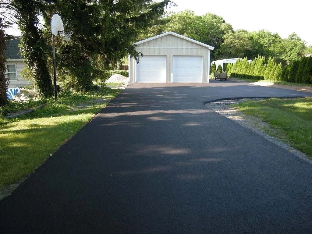 Freshly paved driveway leading to a white double garage beside a green lawn and trees.