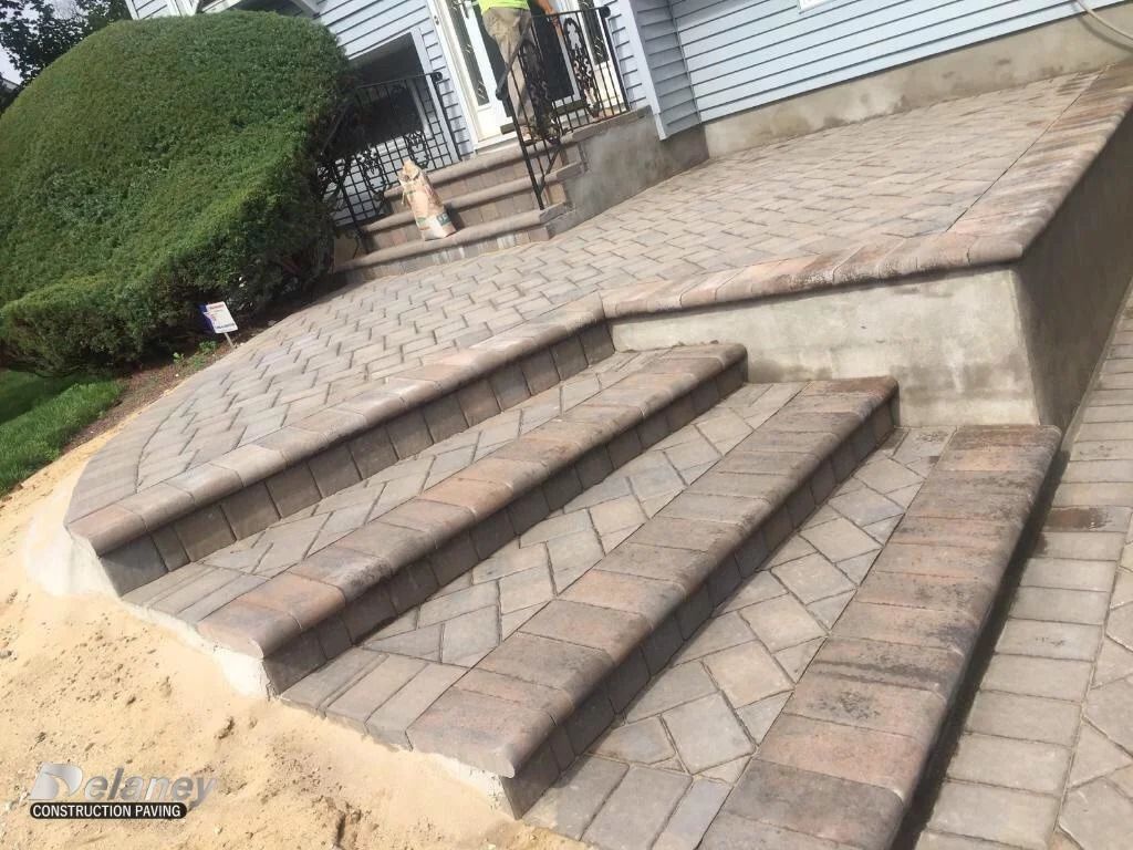 Concrete steps leading to a raised tiled platform beside a sidewalk and trimmed shrub.