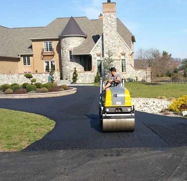 Person on a yellow roller compactor paving a driveway in front of a stone house