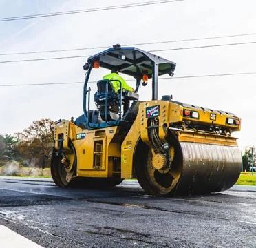 Yellow road roller compacting fresh asphalt on a road, with a worker in a high-vis vest under the canopy.