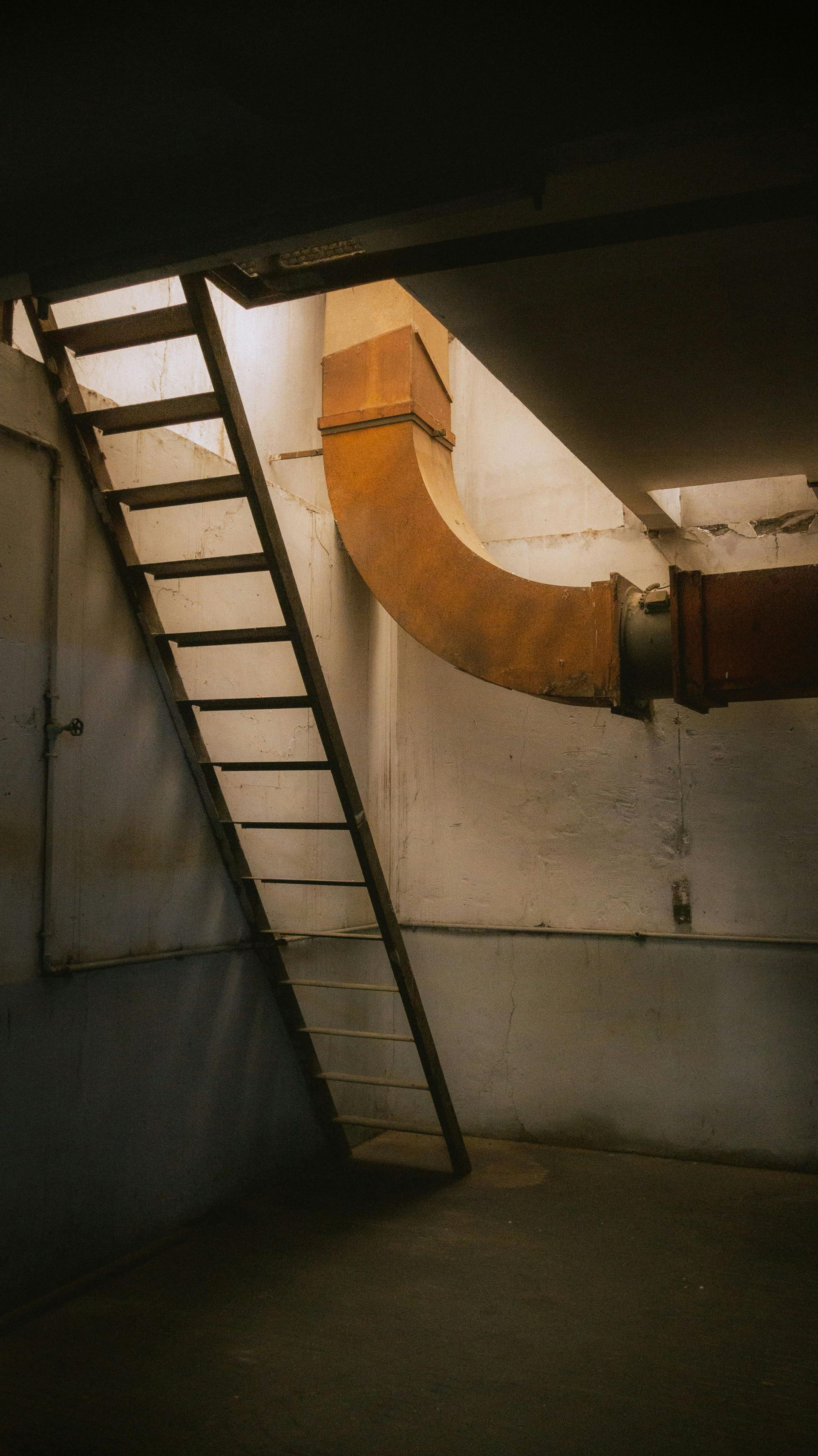 Metal ladder leaning against a concrete wall in a dim industrial basement, with an orange ventilation duct overhead