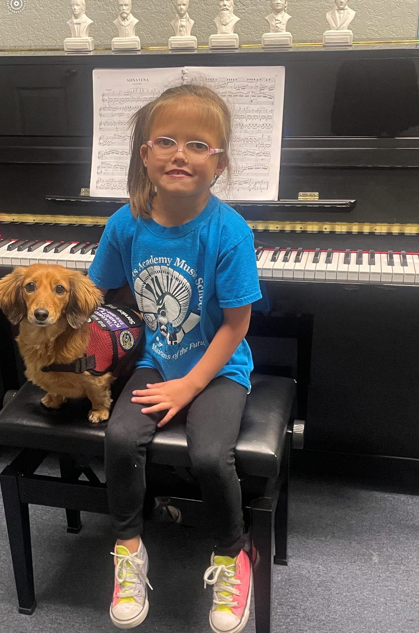 A little girl is sitting on a bench with a dog in front of a piano.