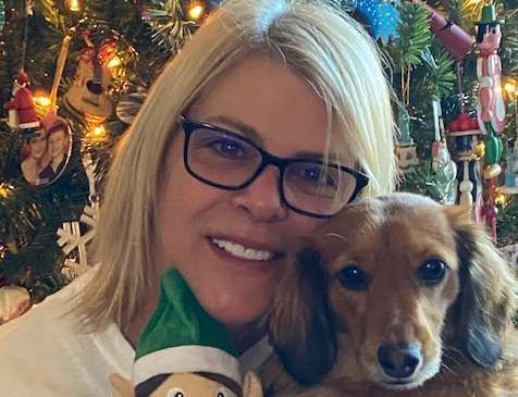 A woman is holding a stuffed elf and a dog in front of a christmas tree.