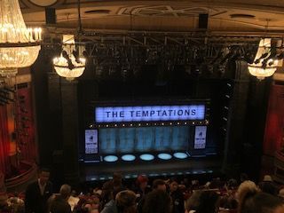 A group of people are sitting in front of a stage with a sign that says the temptations.