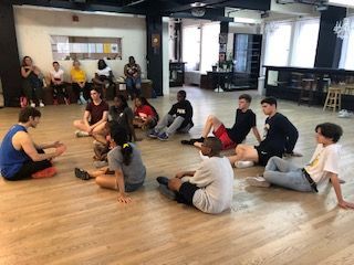 A group of people are sitting on the floor in a circle in a dance studio.