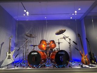 A drum set and guitars are displayed in a glass case
