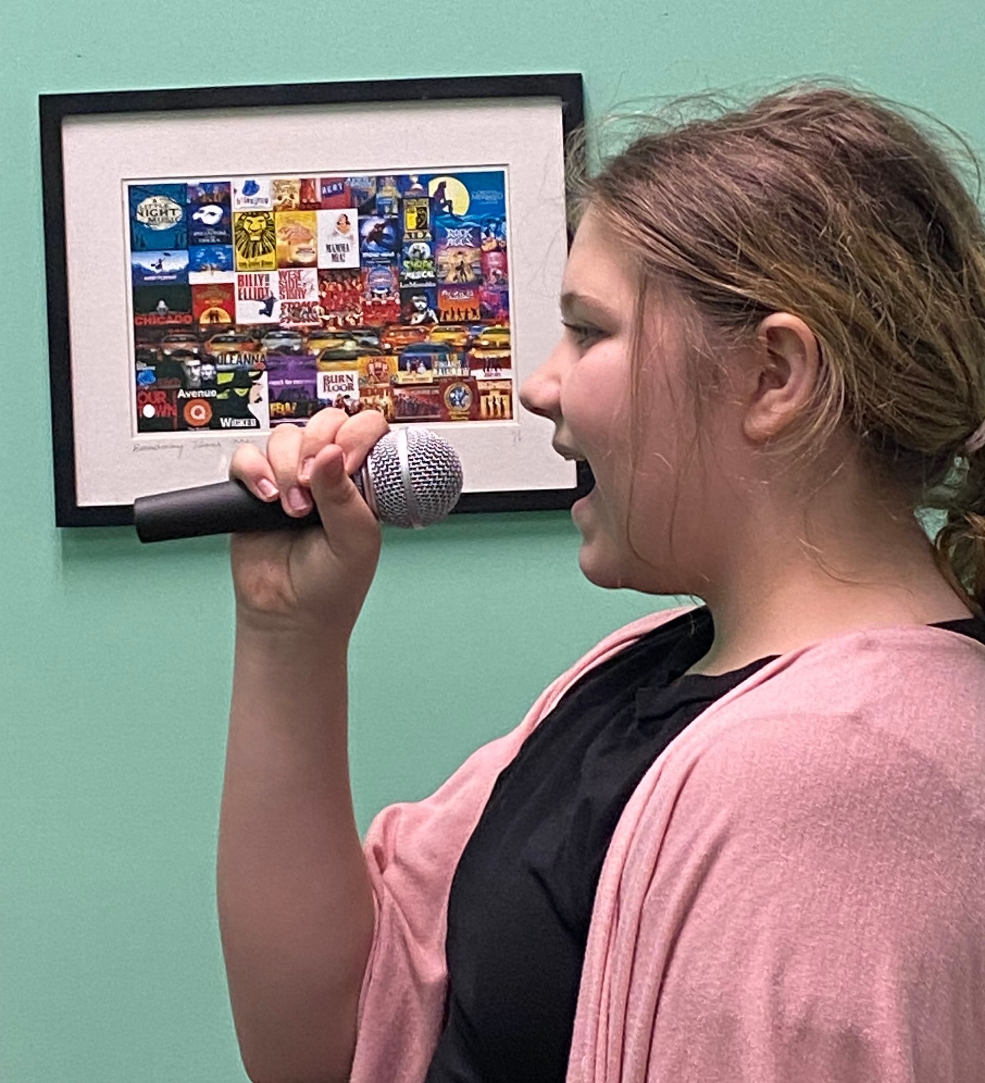 A young girl is singing into a microphone in front of a picture on a wall.