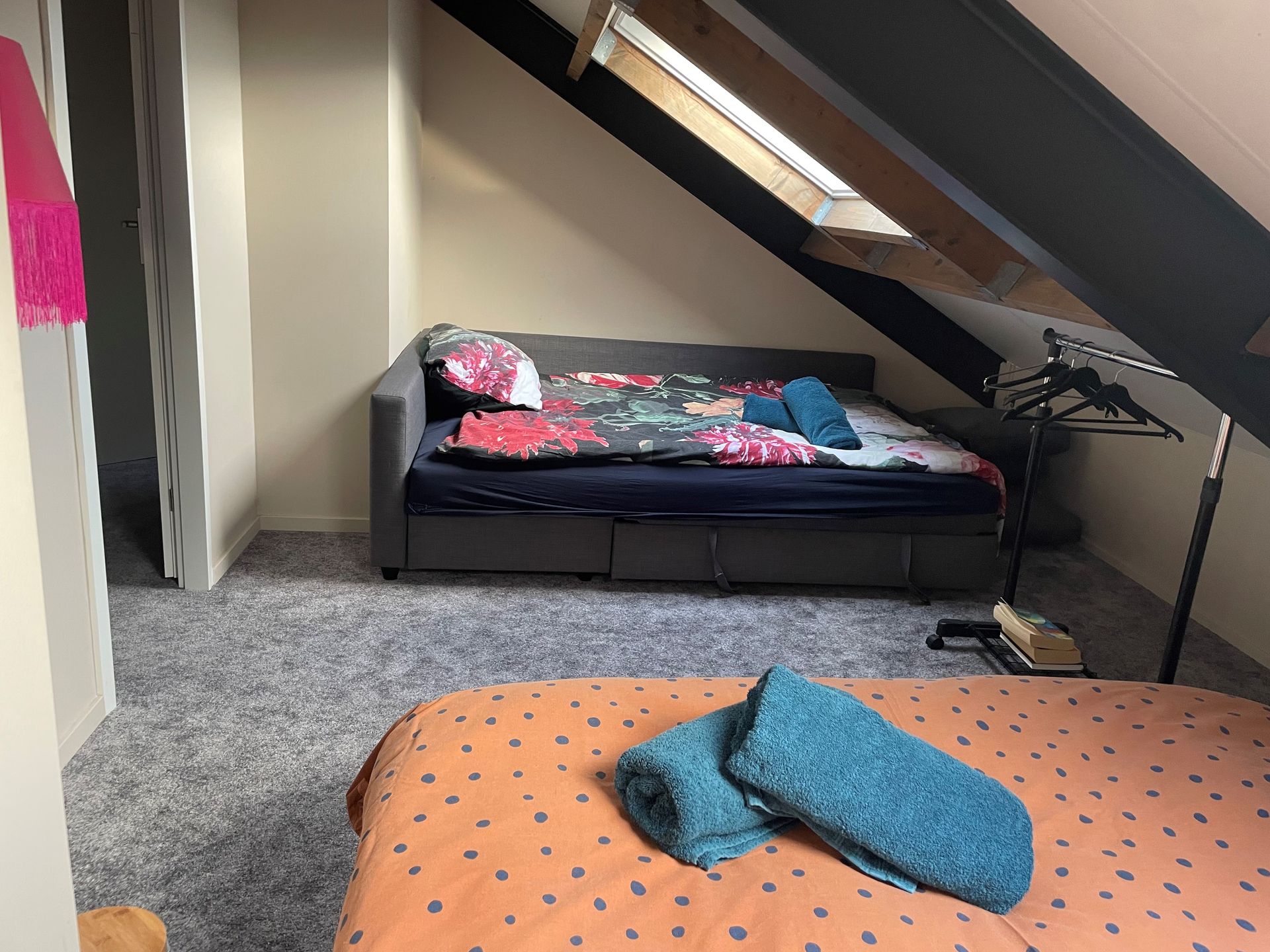 Slaapkamer met twee bedden, een grijs en een oranje. Op een van de bedden liggen handdoeken. Vloerbedekking, schuin plafond.