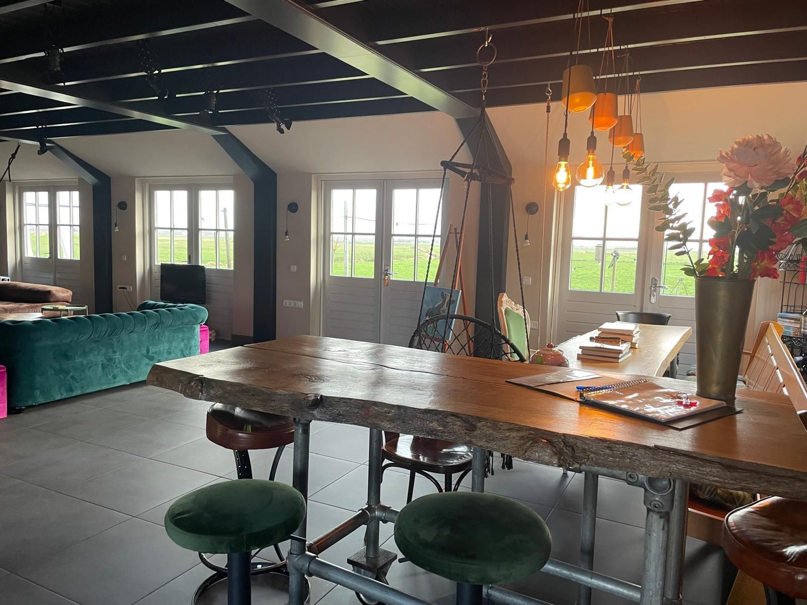 Open woonruimte met een grote houten tafel en barkrukken, een groene fluwelen bank en grote ramen.