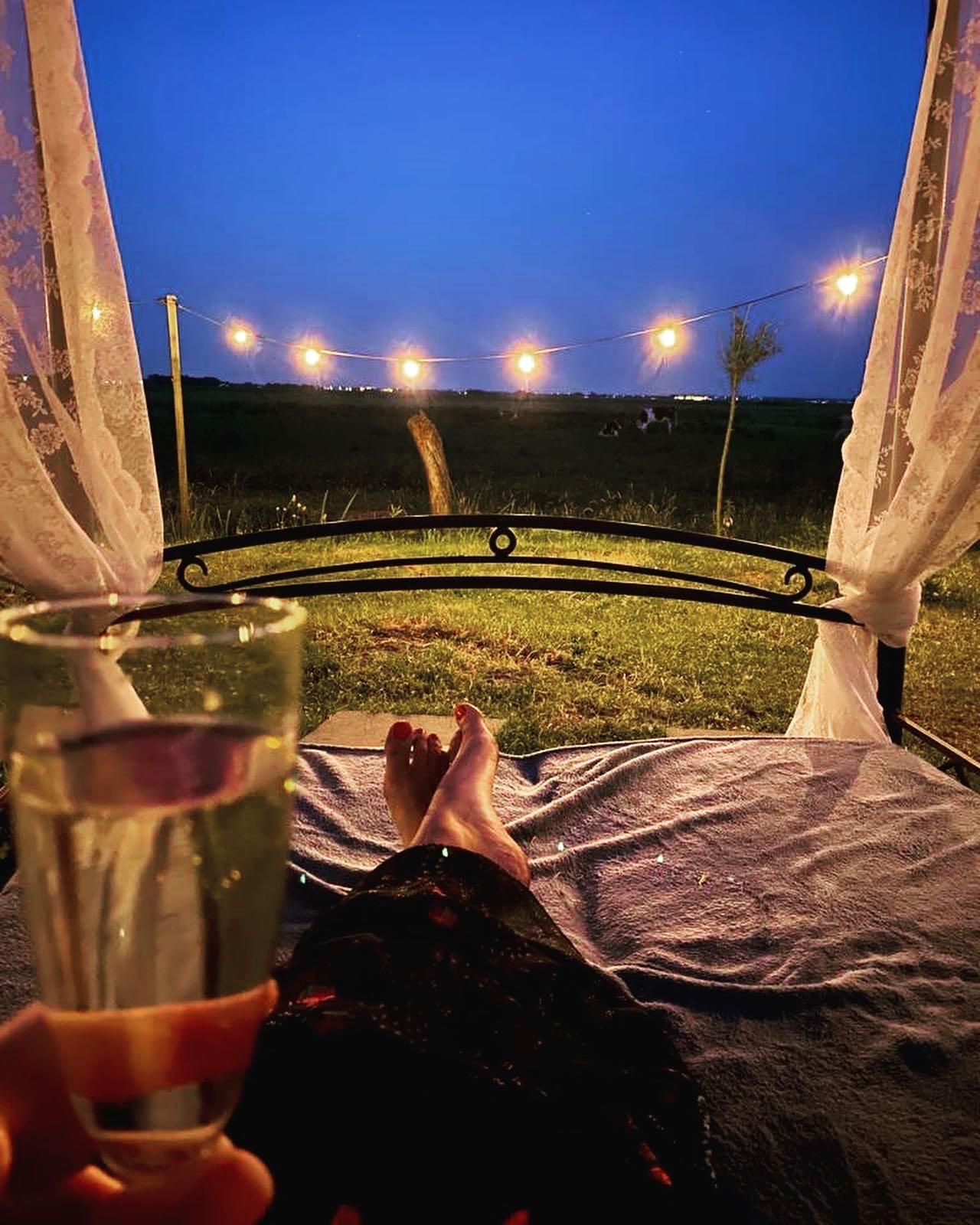 Iemand ligt languit op bed met een drankje en kijkt uit over een veld in de schemering; boven het hoofd hangen lichtslingers.