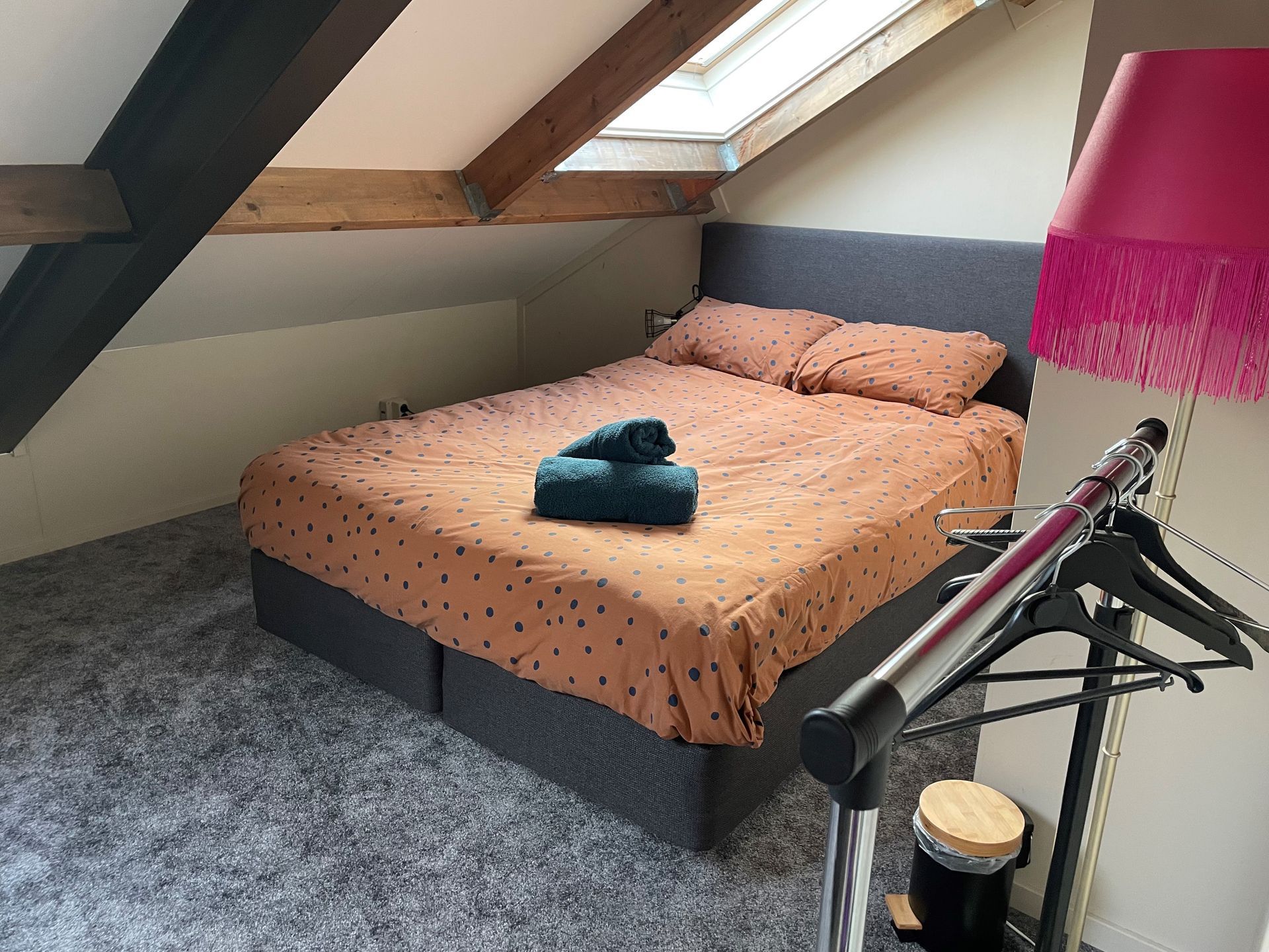 Slaapkamer op zolder met een tweepersoonsbed, grijs tapijt en een roze lamp met franjes.