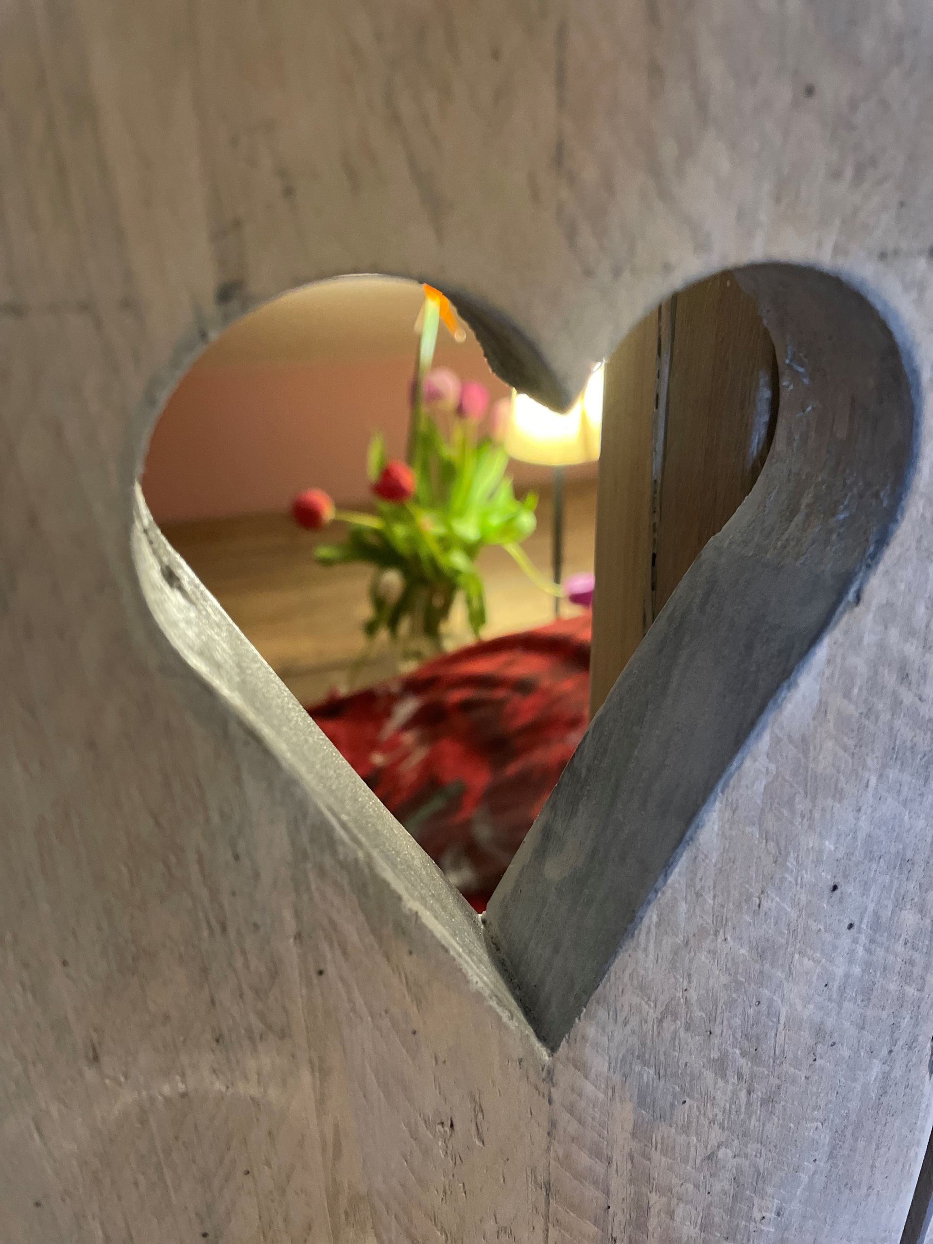 Een hartvormige uitsparing in een houten oppervlak omlijst een wazig beeld van bloemen en een rode deken in een kamer.