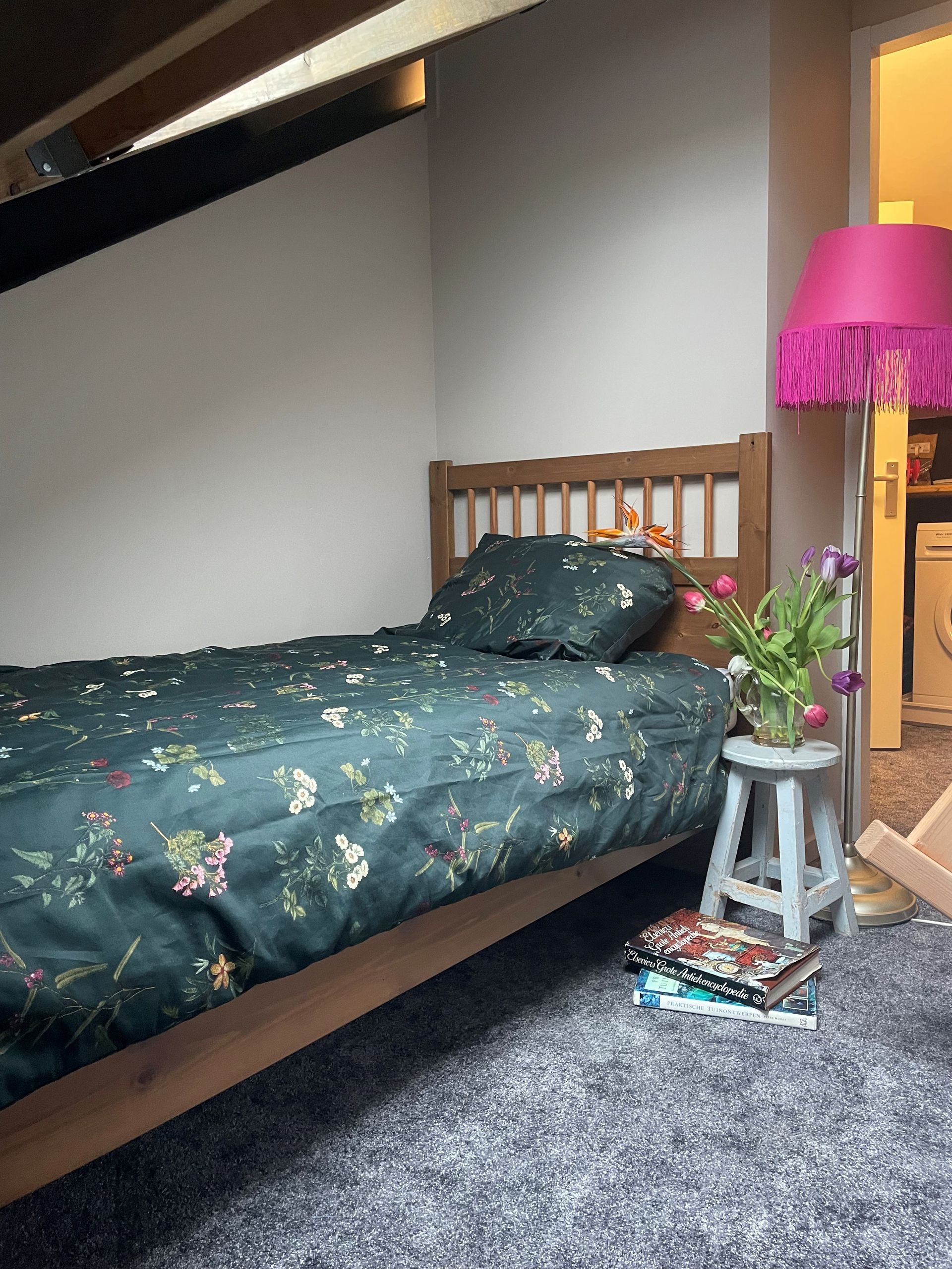 Slaapkamer met een houten bed, beddengoed met bloemenprint, een roze lamp met franjes en een klein krukje met bloemen.