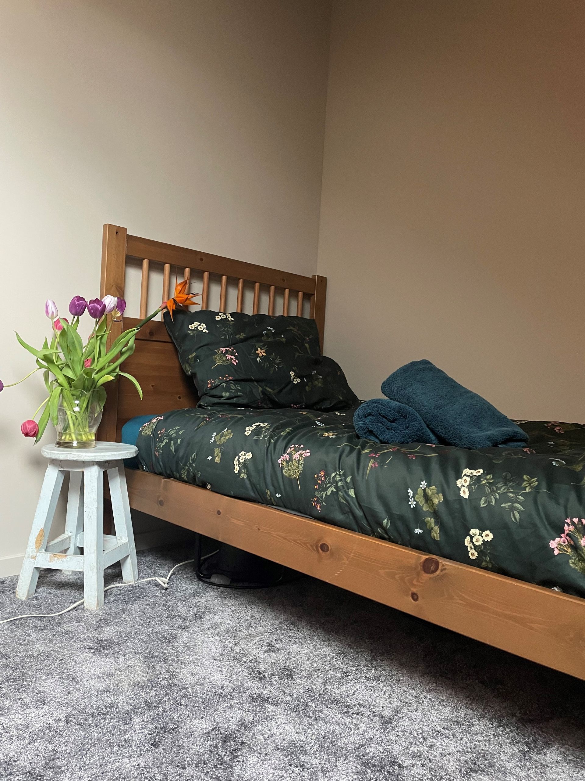 Slaapkamer met houten bed en beddengoed met bloemenprint, bloemen op een krukje en een zacht vloerkleed.
