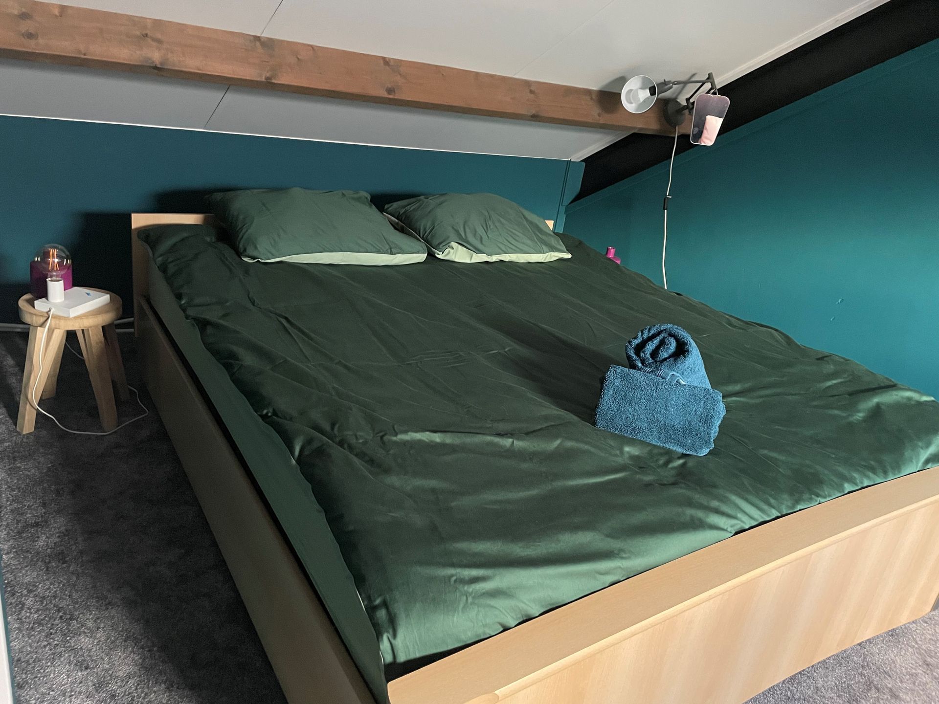 Slaapkamer met een bed opgemaakt met groen beddengoed, een klein krukje en een turquoise muur.