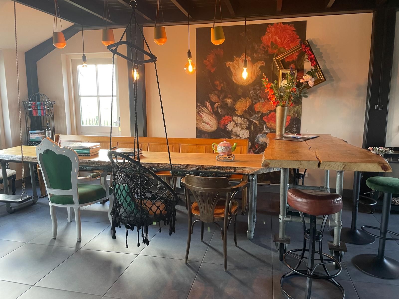 Eclectische eetkamer met houten tafel, hangstoel, bloemstukken en diverse stoelen.