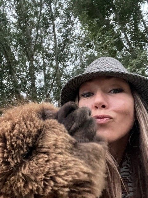 Een vrouw tuit haar lippen naast een bruin, harig dier in de buitenlucht. Beiden kijken onder een hoed naar de camera, voor een boom.
