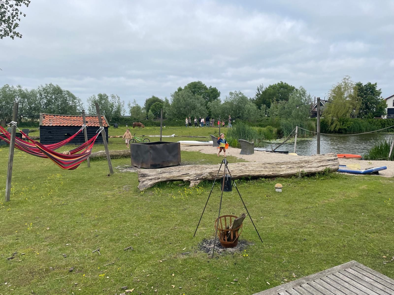 Grasrijk park met vuurplaats, hangmat en waterkant; mensen in de verte.