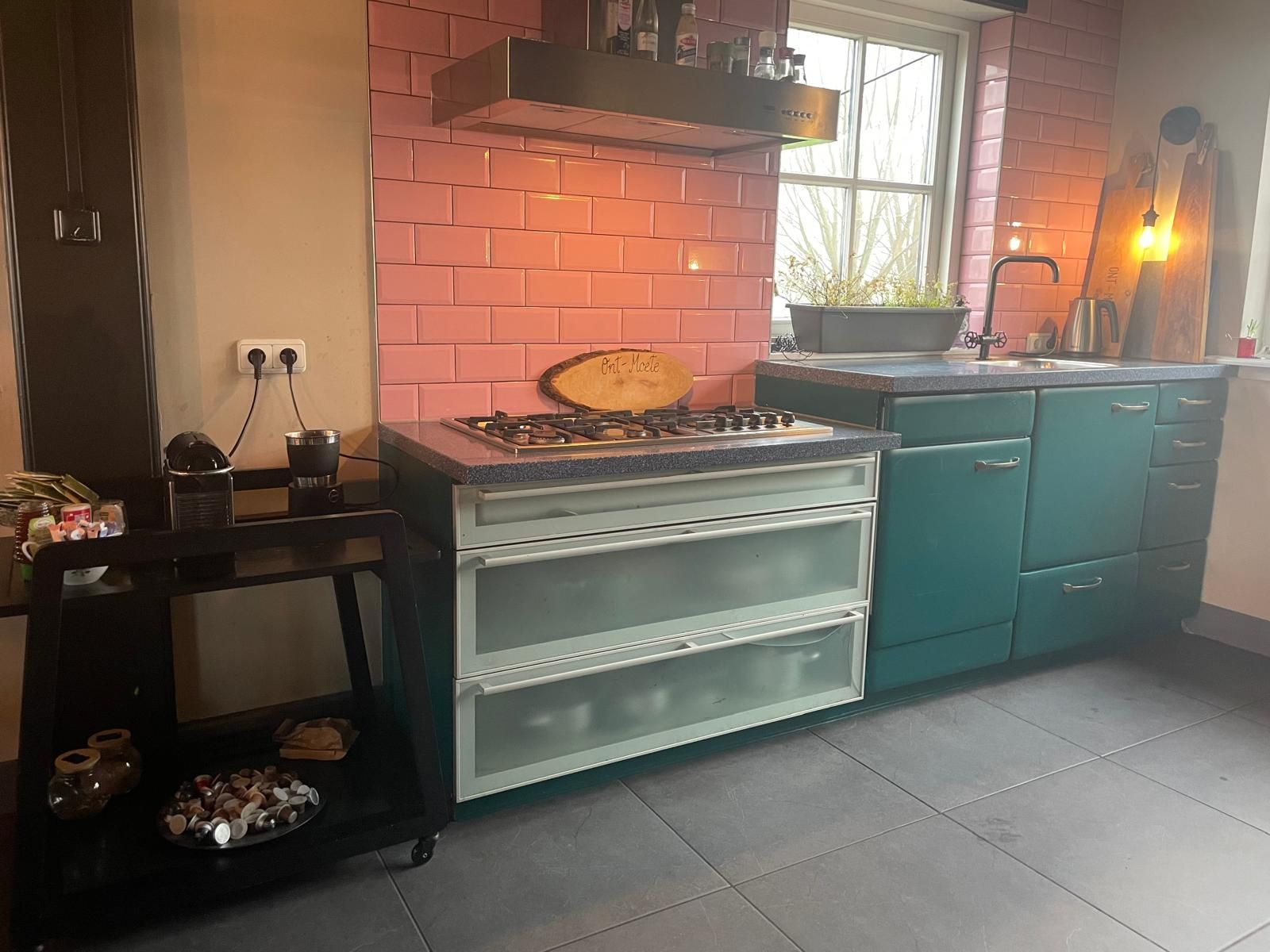 Keuken met turquoise kastjes, roze achterwand, gasfornuis en zwarte trolley.