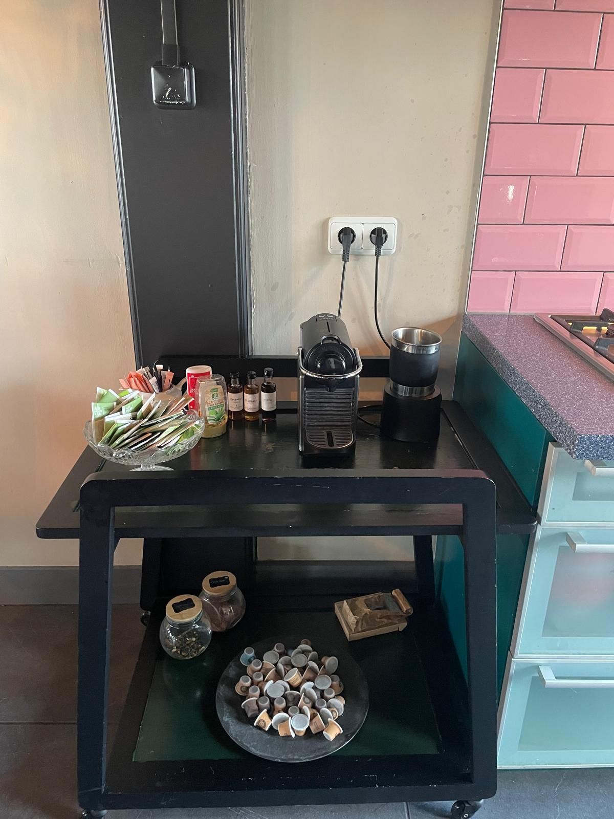 Zwarte trolley met koffiezetapparaat, thee en snacks; vlakbij de muur met stopcontact en opbergruimte.