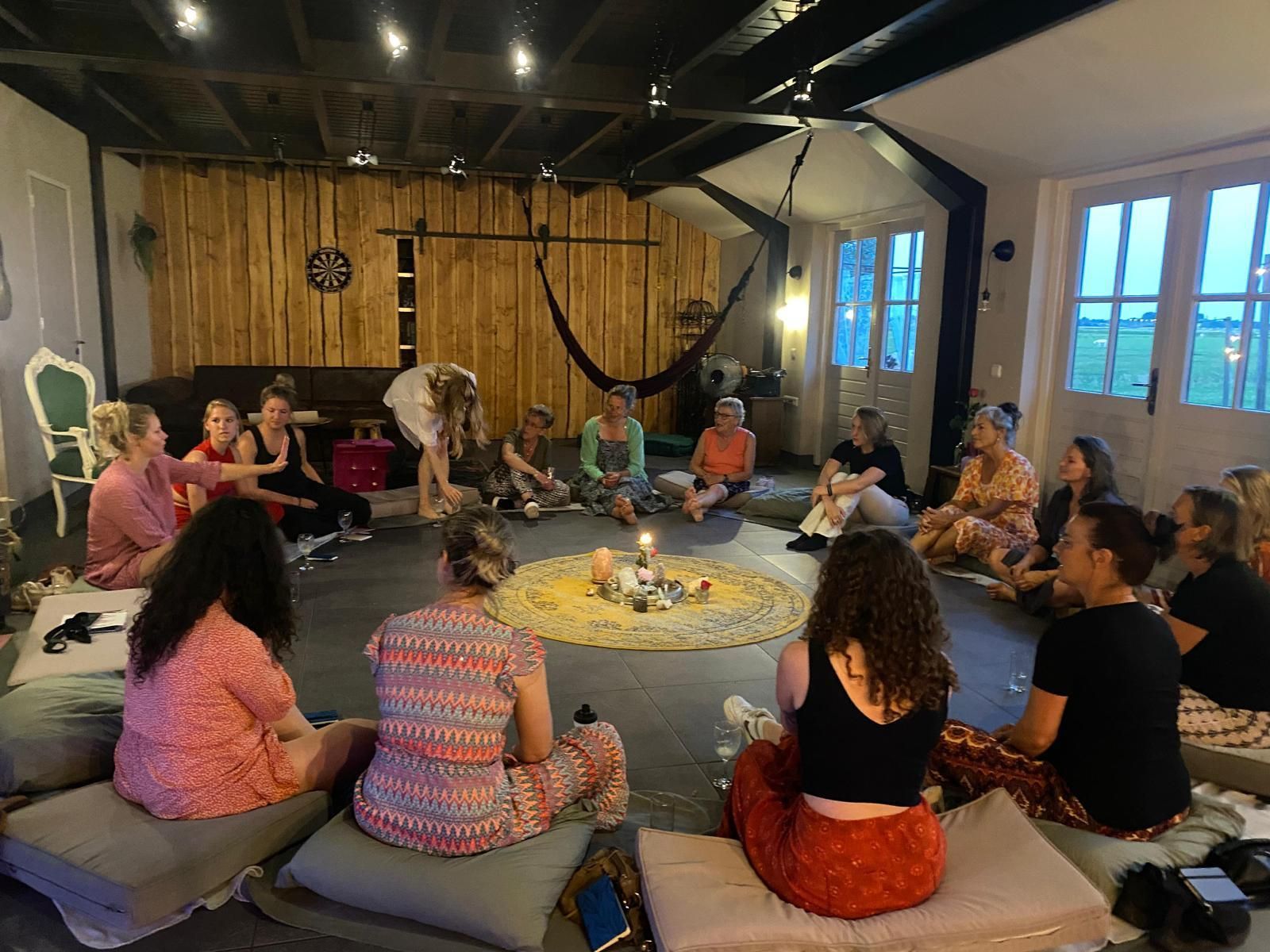 Mensen zitten in een kring rond kaarsen op kussens. Binnenruimte met houten wanden en ramen.