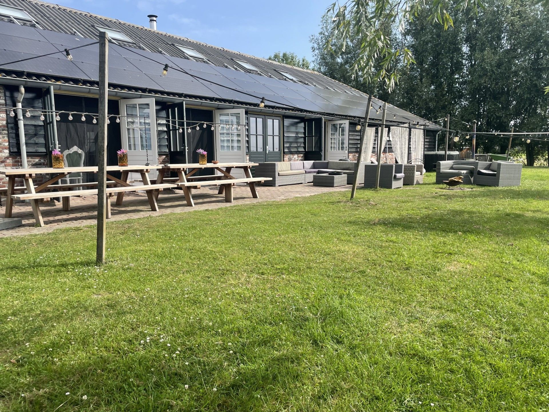Buitenpatio met picknicktafels, loungebanken en een grasveld voor een gebouw met witte deuren.