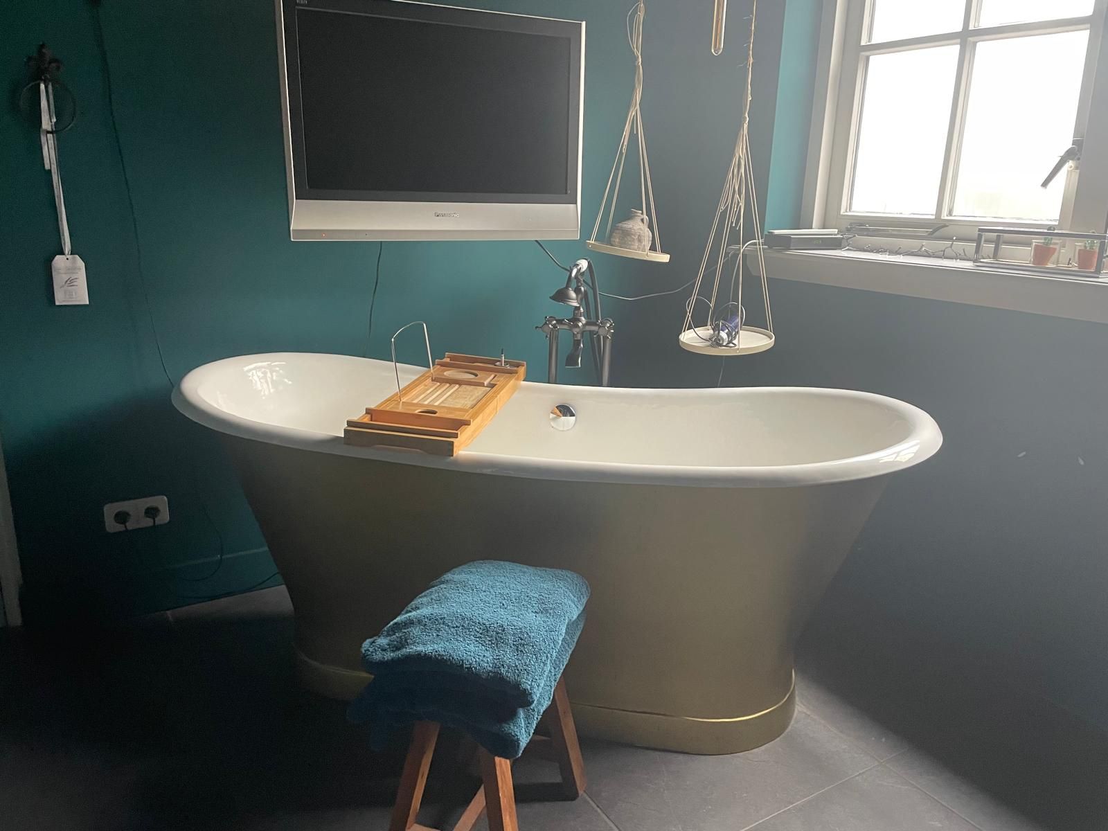 Vrijstaande gouden badkuip in een turquoisekleurige badkamer met een aan de muur gemonteerde tv en macramé plantenhouders.