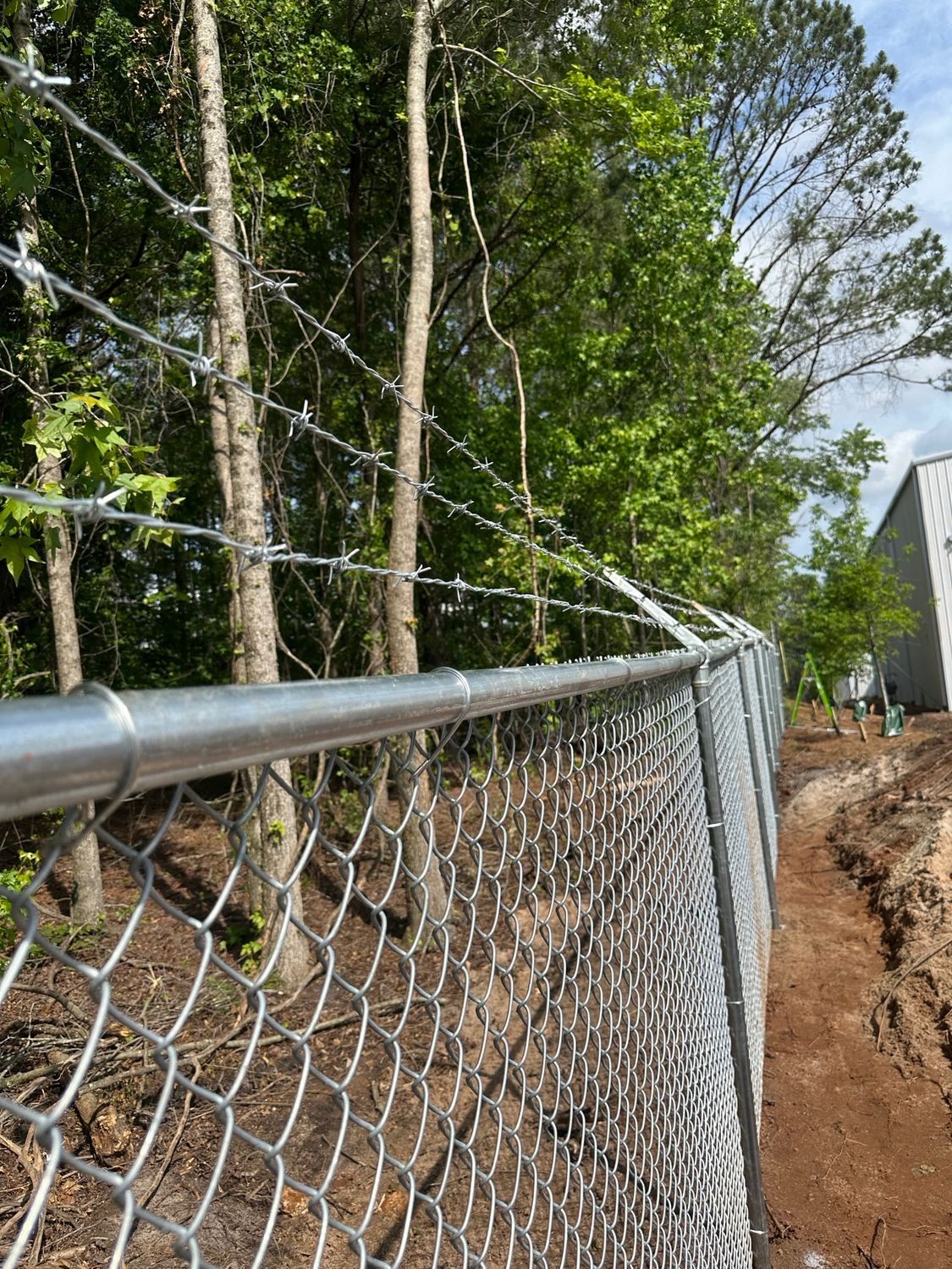 Chain link fence with barbed wire topper, near trees and a building.