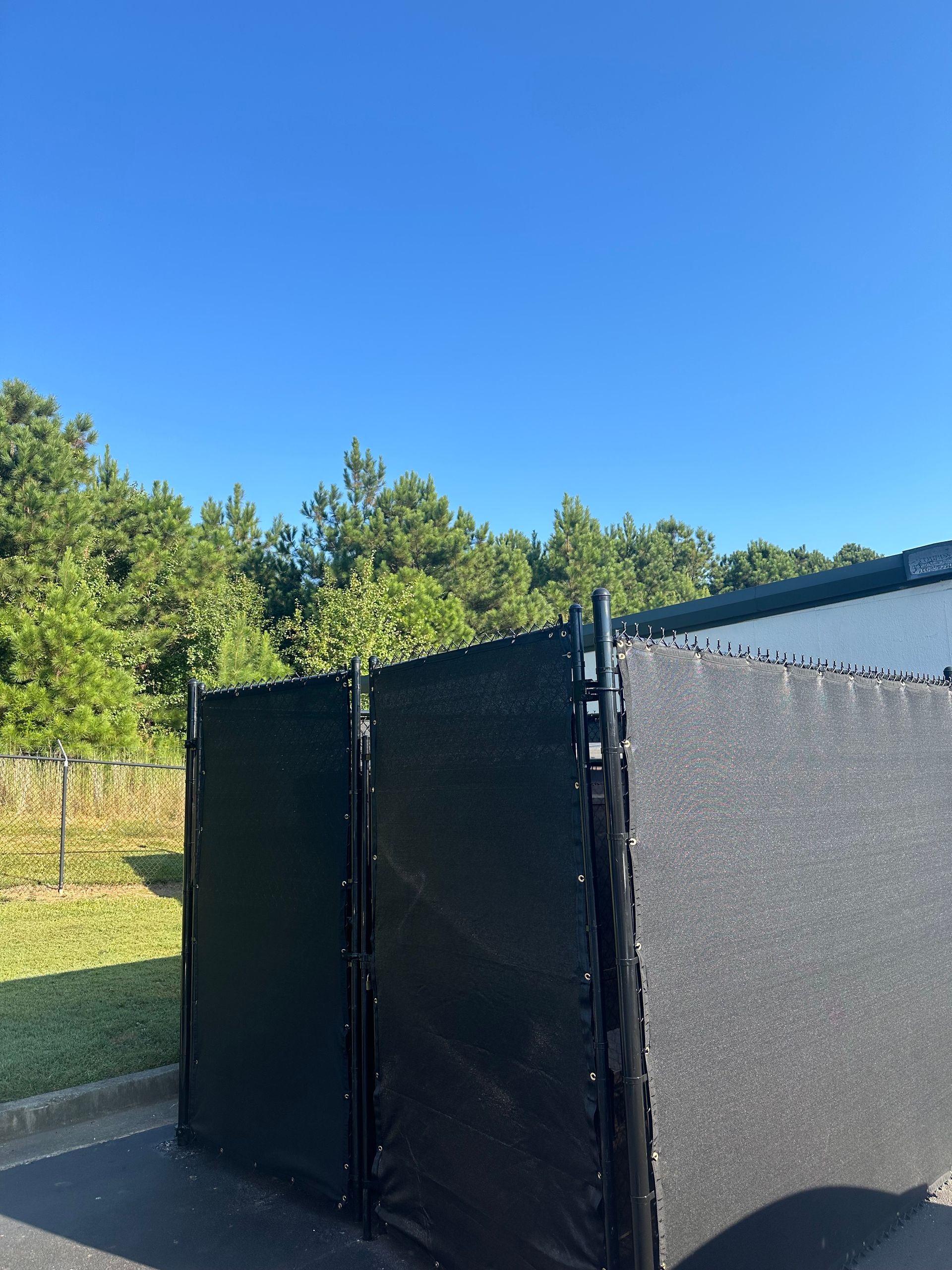 Black and gray privacy fence against a backdrop of trees and a bright blue sky.