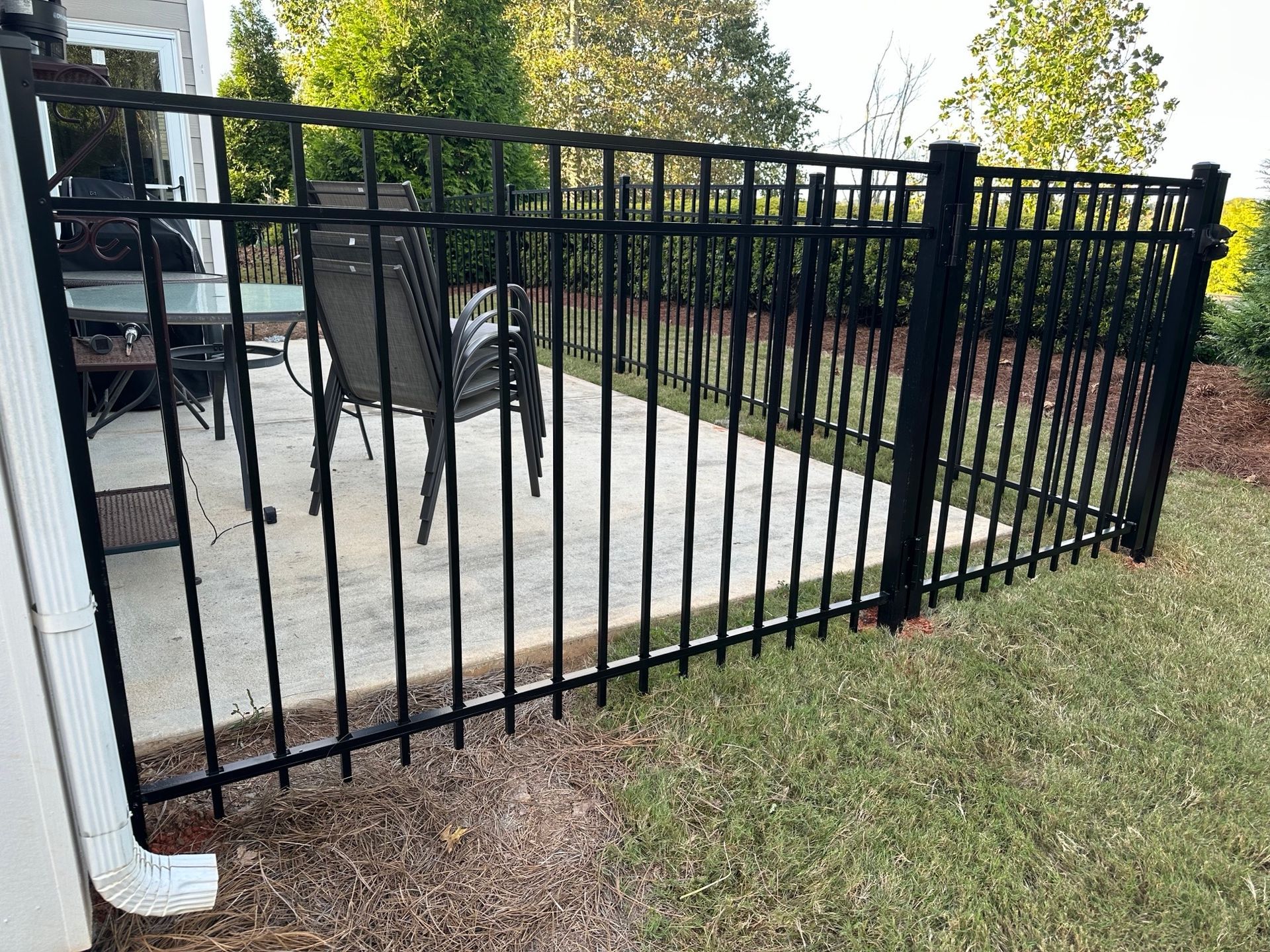 Black metal fence encloses a concrete patio with a table and chairs on a grassy yard.
