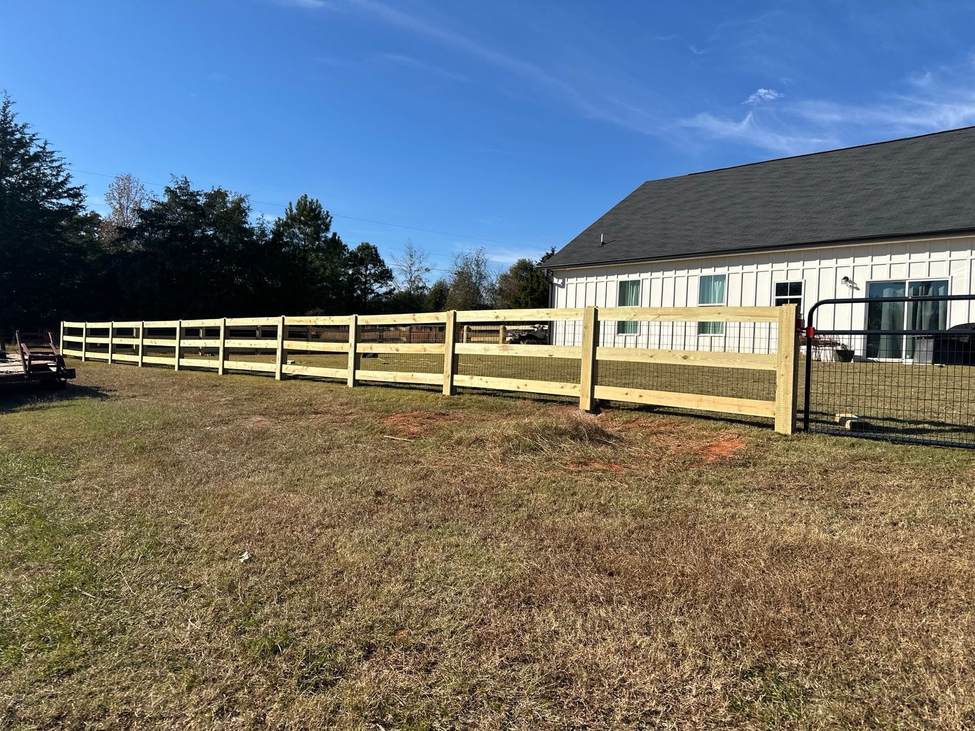Wooden split-rail fence surrounds a grassy yard, beside a house with a dark roof and a clear blue sky.
