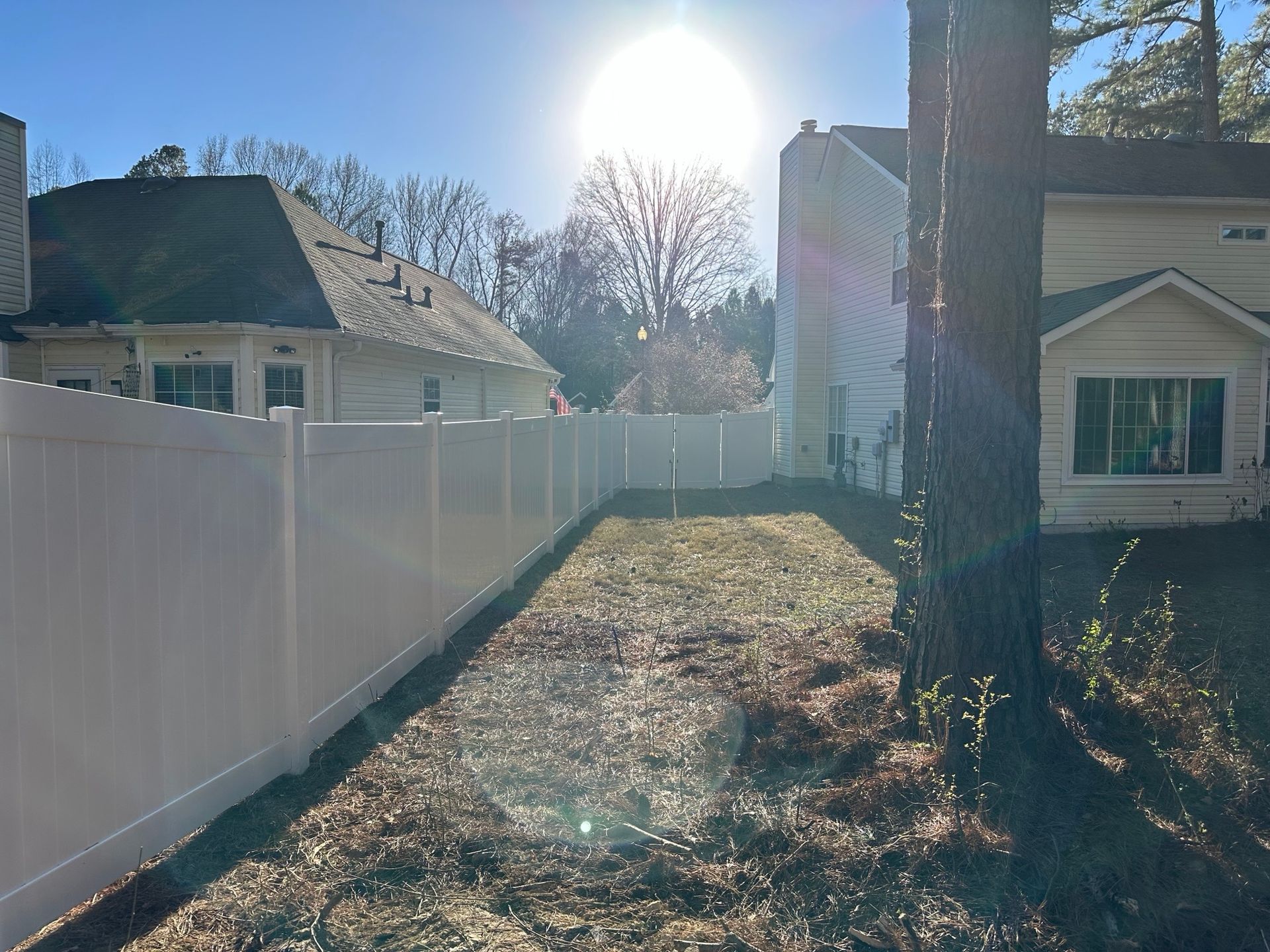 White fence runs alongside a grassy yard. Houses and trees are in the background, sun shining brightly.